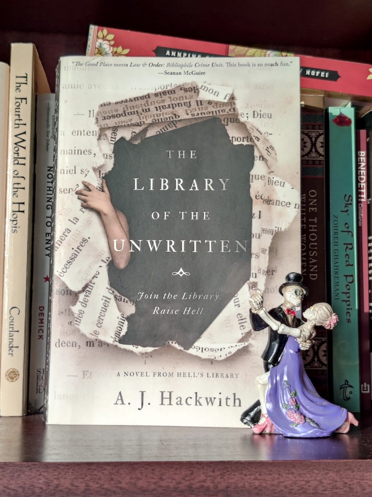 Image of a book on a shelf. Other books of various sizes are in the background. The book cover is in sepia, black, and white colors and has written at the top in black "The Good Place meets Law & Order: Bibliophile Crime Unit. This book is so much fun. -- Seanan McGuire. The image on the cover is of an arm coming out of the left side of a dark opening of ripped pages from what seems to be a book. The words on the ripped pages are in french. The title "The Library of the Unwritten" is in the dark opening where the arm is coming out of in white letters. Underneath the title it says "Join the Library. Raise Hell. Underneath the opening it says "A novel from Hell's Library" and under that "A. J. Hackwith"